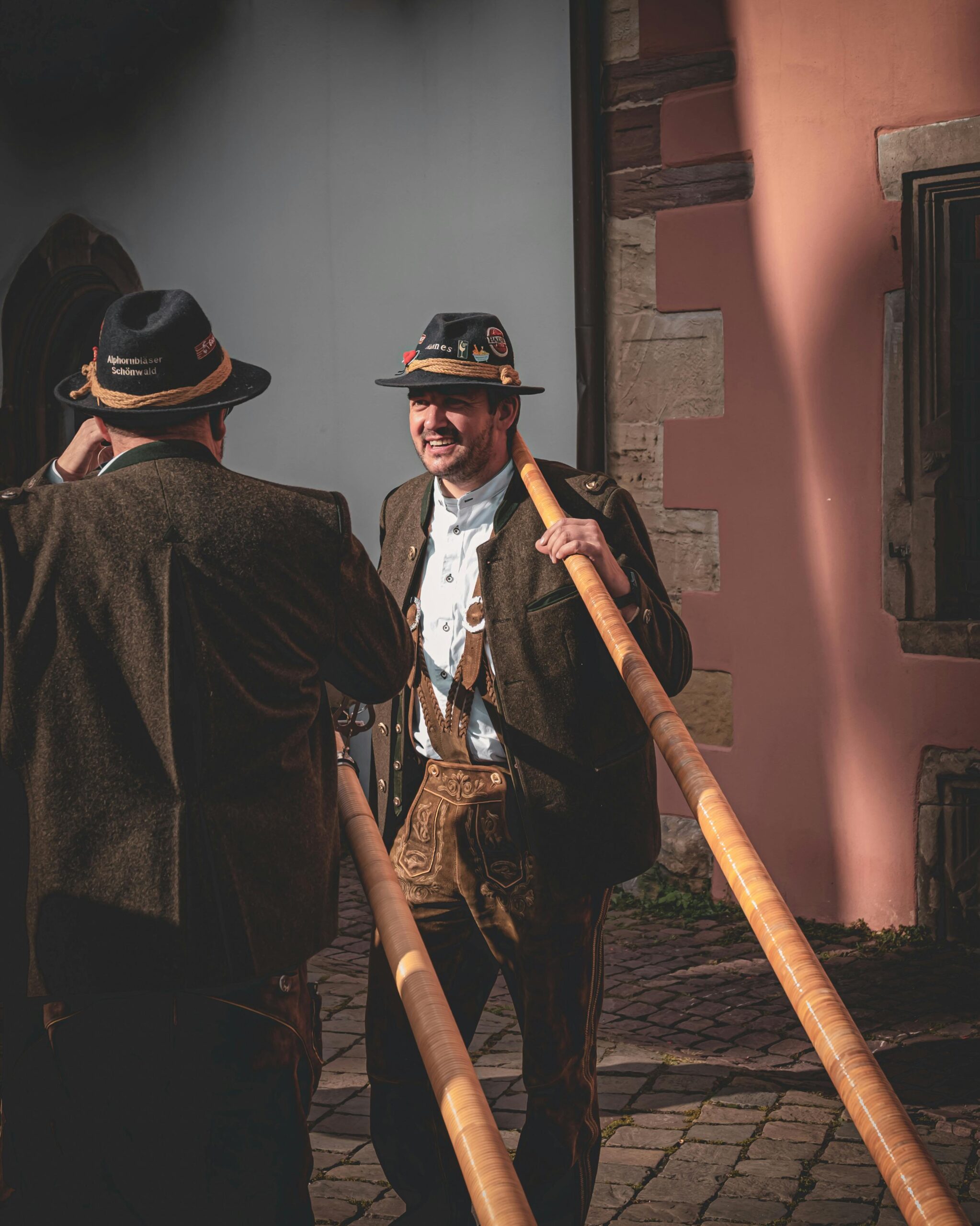 Two male Bavarian musicians play alphorns in traditional attire outdoors.